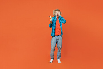 Fototapeta na wymiar Full body sad astonished stupefied young blond caucasian man wear blue shirt orange t-shirt hold head use mobile cell phone isolated on plain red background studio portrait. People lifestyle concept.