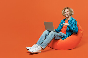 Full body happy fun young blond IT man wear blue shirt orange t-shirt sit in bag chair hold use...
