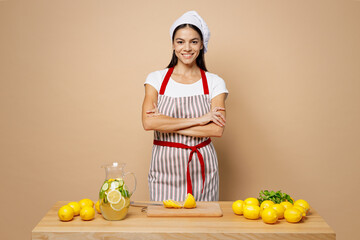 Young confident housewife housekeeper chef latin woman wears apron toque hat work at table kitchenware cutting lemons for lemonade isolated on plain pastel light beige background. Process cook food.