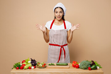 Young fun surprised housewife housekeeper chef cook latin woman wear apron toque chefs hat work at table kitchenware spread hands isolated on plain pastel light beige background. Process cooking food.