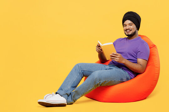 Full body devotee Sikh Indian man ties his traditional turban dastar wearing purple t-shirt sit in bag chair reading book in leisure fre spare time isolated on plain yellow background studio portrait.