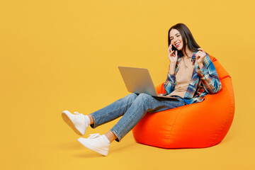 Full body young woman wear blue shirt beige t-shirt sit in bag chair hold use work on laptop pc computer talk on mobile cell phone isolated on plain yellow background studio. People lifestyle concept.