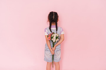 Little girl in denim overalls holding bouquet of roses behind her back against pink background. mother's day
