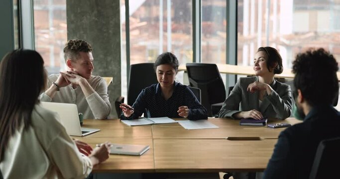 Confident Indian team leader speaking to employees at corporate meeting, sitting at conference table, giving instruction, sharing ideas. Workgroup discussing ideas for project in office, laughing