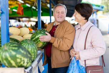 Aged man and woman customers buying watermelon in open-air market