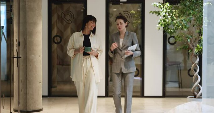 Confident friendly businesswomen talking in office hall, walking in corridor. Corporate friends chatting on work break, speaking, listening, smiling, laughing, enjoying friendship, networking