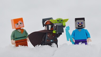 Fototapeta premium LEGO figures of Minecraft Steve and Alex standing together with LEGO Star Wars Mandalorian with baby Yoda, also known as Grogu on his left arm, in deep winter snow.