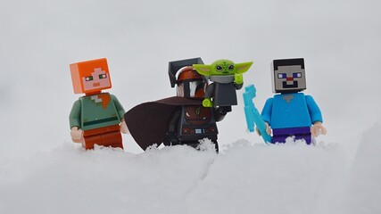 Naklejka premium LEGO figures of Minecraft Steve and Alex standing around LEGO Star Wars Mandalorian with baby Yoda, also known as Grogu on his left arm, in deep winter snow.