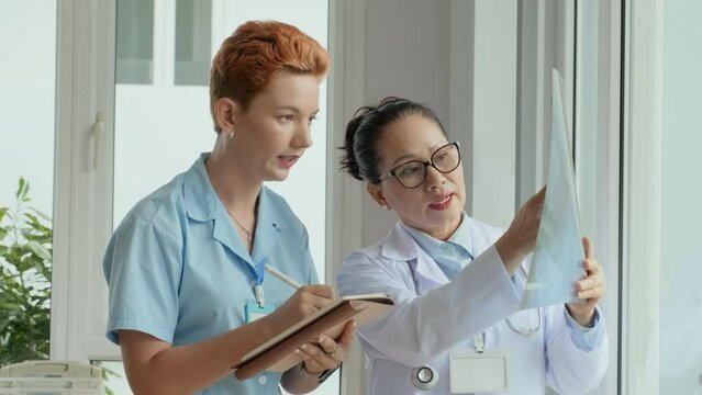 Young female clinician in blue uniform asking questions to experienced radiologist and writing down her answers in notepad during discussion of patient x-ray results