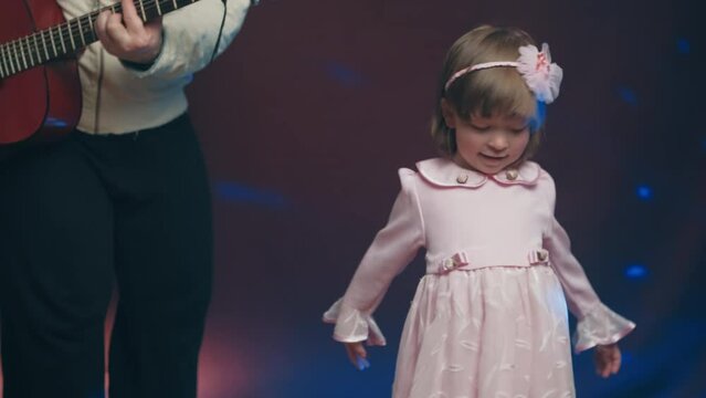 Little girl on stage in pink vintage dress, she dances, her father plays acoustic guitar. Color music is shining. Performance on stage. Early development of children