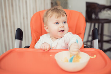Fototapeta premium Cheerful happy child eats food by himself with a spoon. Portrait of a happy boy in a high chair for feeding.