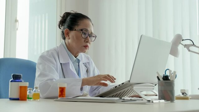 Mature confident clinician in lab coat and eyeglasses sitting in front of laptop, looking through medical data in document, then taking bottle of pills and consulting online patients