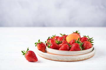 Fresh strawberries and apricot in two ceramic plates on a white wooden table and a gray background....