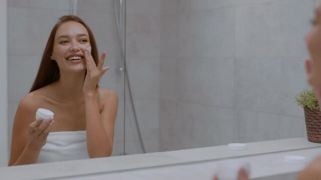 Morning beauty procedures. Young happy smiling woman applying pampering cream on face, looking at mirror at bathroom