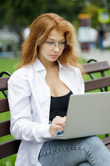 Young red-hair female student using her laptop sitting on the bench in park.