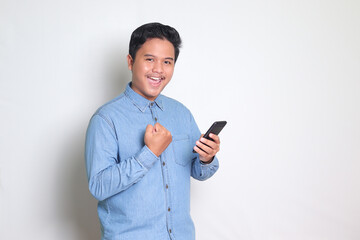 Portrait of Asian man in blue shirt raising his fist, celebrating success, winning game on his mobile phone. Isolated image on white background