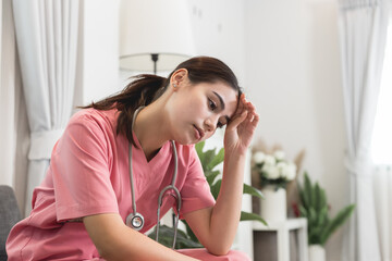 Fototapeta na wymiar Upset female nurse sitting on a couch at home. Healthcare worker having headache. Doctors face heavy levels of stress. Young Caucasian caregiver in pink scrubs sitting hand touching temple tired face.
