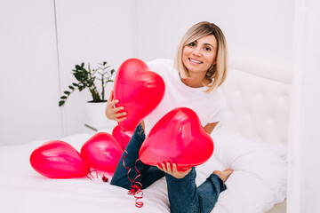 Excited Girl sitting on bed with red hearts shape balloons in white interior. Woman rejoices gift on valentines day. Easy-going girl with blond hair expressing positive emotions in valentines day.