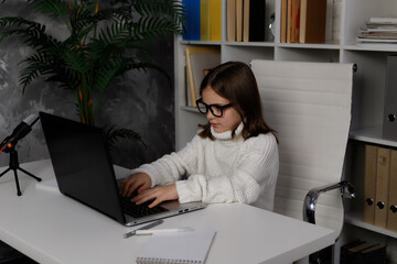 Little school girl in glasses typing, using laptop, working from home. Kid doing homework. Caucasian brunette child at white table. Concept of online education, e learning, technologies