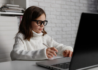 Little school girl in glasses using laptop at home. Kid doing homework. Caucasian brunette child at white table. Concept of online education, e learning, technologies