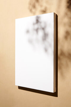 White canvas, blank picture mockup hanging on beige wall with dark shadows of leaves. Poster mockup, empty canvas with shadows of plant, side view