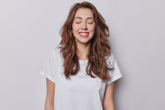 Waist up shot of pleased long haired woman keeps eyes closed smiles toothily chuckles as hears something funny dressed in casual basic t shirt isolated over white background. People emotions concept