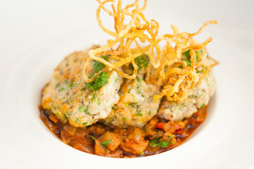 Close-up of three crab cakes on a bed of vegetable stew in red sauce. Meal topped with pesto and abstract fried calamari. Modern food plating. Chef's special on white plate. Horizontal background.
