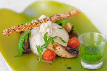 Close-up of burrata cheese salad with mushrooms, cherry tomatoes, arugula, a bread stick with sesame seeds, pesto sauce. Meal on a neon green plate. Modern food plating. Horizontal lunch background.