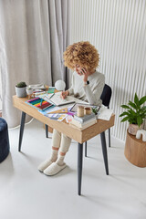 Fototapeta na wymiar Indoor shot of professional curly haired female interior designer plans design of future apartment asks for advice in colleague via smartphone poses at table makes room drawings works from home