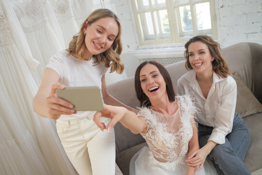 Women taking photographs of a female friend trying on wedding dress. Women in wedding dress fitting room.