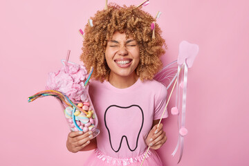 Horizontal shot of extremely happy curly haired woman holds glass of sweet colorful candies and...