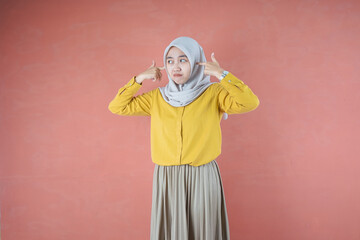 Beautiful Asian woman in yellow shirt and hijab covering ears with hands on brown background