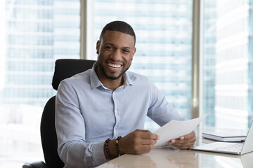 Excited happy millennial African business man holding legal documents at work office table, looking...