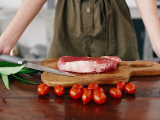 Woman with knife in hand cutting fresh steak meat for roasting in kitchen with salt pepper and other spices on table, red cherry tomatoes and herbs, preparing dinner.