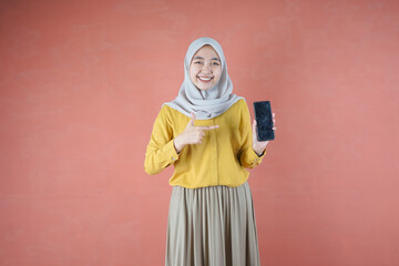 Fototapeta premium Beautiful Asian woman in yellow shirt and hijab smiling cheerful has smartphone with black screen in hand, pointing with index finger at product on brown background