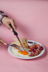 hand with knife and fork cuts breakfast, scrambled eggs with tomatoes and toast, plain background, side view
