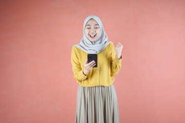 Beautiful Asian woman in yellow shirt and hijab smiling cheerful using mobile phone gesturing yes with raised fist on brown background