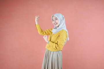 Beautiful Asian woman in yellow shirt and hijab smiling cheerful winner gesture celebrate clenching fists say yes on brown background