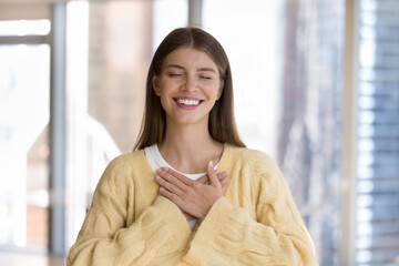 Happy pretty young woman applying hands at chest with gratitude, posing with closed eyes and toothy...