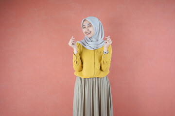 Fototapeta na wymiar Beautiful Asian woman in yellow shirt and hijab smiling cheerfully showing Korean heart with two fingers crossed, express joy and positivity over brown background