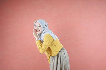 Beautiful Asian woman in yellow shirt and hijab smiling cheerfully whispering over brown background