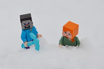Fototapeta premium LEGO Minecraft figures of Steve and Alex climbing on snowy hill, winter cloudy sunshine. 