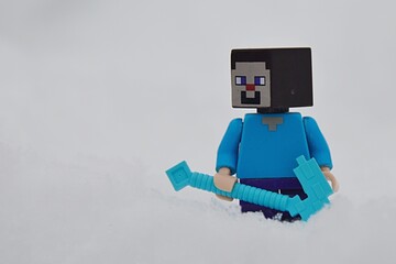 Fototapeta premium LEGO Minecraft smiling figure of main character Steve standing with his diamond pickaxe in deep snow.