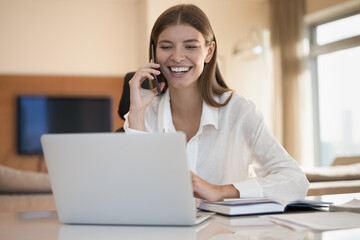 Cheerful freelancer woman enjoying job communication, sitting at home office table, looking at laptop, talking on mobile phone, smiling, laughing, discussing career success