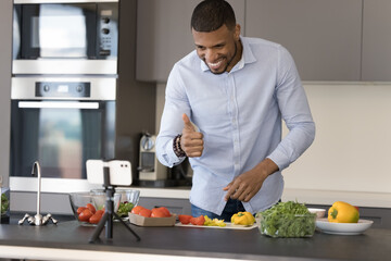 Cheerful successful chef blogger guy shooting cooking masterclass, video lesson, promoting healthy eating, preparing salad at smartphone web camera, showing like thumb up to audience