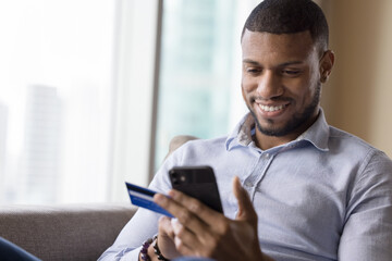 Happy millennial man using credit card, ecommerce app on mobile phone for payment, online order, shopping, buying, sitting on home couch, typing, smiling, laughing - obrazy, fototapety, plakaty