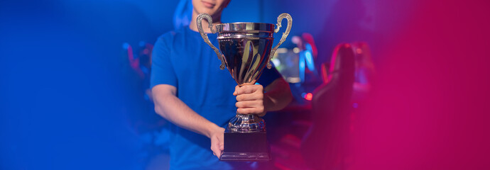 Concept banner cyber gaming esport. Winner gamer young man holding cup trophy prize of victors online video games tournament. Soft focus, neon color © Parilov