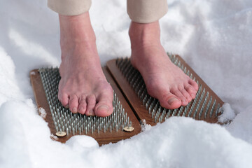 A woman practicing yoga therapy stands on a board with nails against a background of snow.