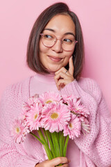 Charming dark haired young Asian woman smiles gently looks pensively aside keeps hand on cheek wears transparent eyeglasses and knitted jumper holds bunch of gerbera flowers has romantic mood