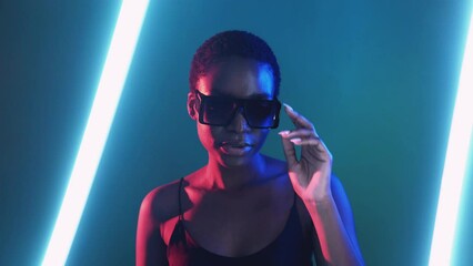 Cyber fashion. Neon eyewear. Futuristic accessories. Confident female model posing in stylish sunglasses in red blue color light isolated on dark night with white LED lamp glow.